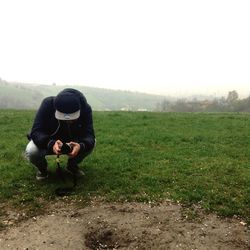 Man photographing while crouching on grassy field against clear sky during foggy weather