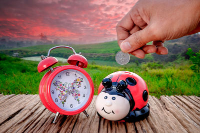Cropped hand inserting coin in piggy bank by alarm clock on table against sky during sunset