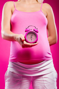 Midsection of pregnant woman holding alarm clock while standing against pink background