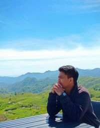 Young man sitting on mountain against sky