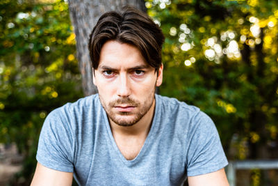 Portrait of serious young man sitting against trees at park