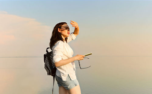 Young woman using phone while standing on sea against sky