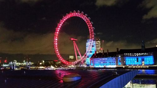 Illuminated ferris wheel by river against sky at night