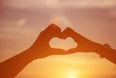 Cropped image of hand holding heart shape against sky during sunset