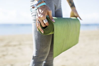 Midsection of woman holding rolled up exercise mat on beach