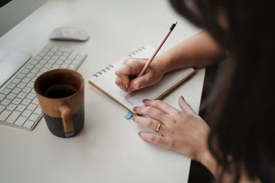 Crop from above view of unrecognizable woman taking notes with pencil while sitting at table with computer and coffee cup in office in paris