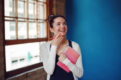 Smiling woman holding book while standing against wall