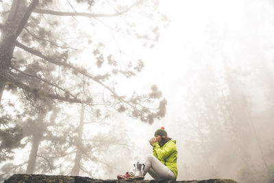 Woman drinking coffee while sitting on retaining wall at forest during foggy weather