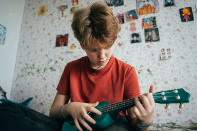 A teenage girl sitting on the bed learns to play the ukulele.