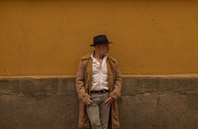 Portrait of adult man in hat and coat against yellow wall on street