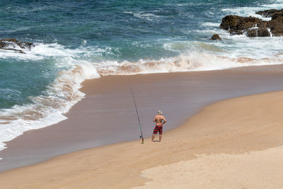 Rear view of shirtless man fishing while standing on shore at beach