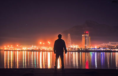 Silhouette man looking at view while standing by sea in illuminated city at night