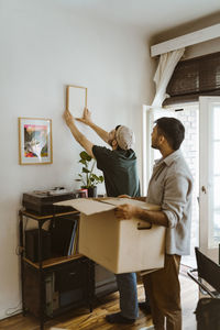 Side view of man with cardboard standing by boyfriend mounting frame on wall at home