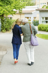 Full length rear view of affectionate female caretaker and senior woman walking on street