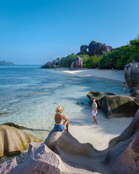 Rear view of woman sitting on rock at beach against clear blue sky