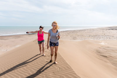 Front view of two women walking on top of a large dune, one of them with arms open to the wind