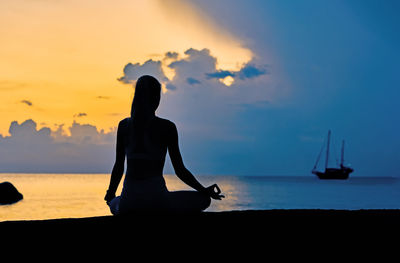 Silhouette of young woman sitting  in meditation on the rock by the sea against sky on sunrise