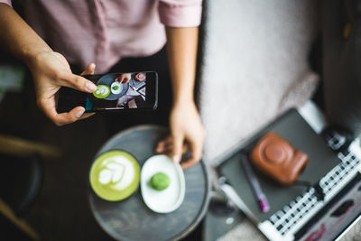 Midsection of female blogger photographing matcha tea and snack through smart phone at creative office