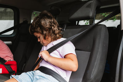 Unemotional girl in casual clothes buckling up while sitting on seat inside of car on back seat of car during road trip