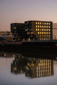 Illuminated building by river against sky at dusk