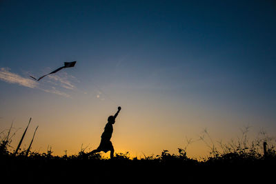 Side view of silhouette boy flying kite on field against sky during sunset