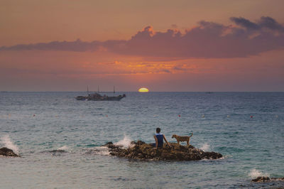 Rear view of man sitting with dog on rock formation in sea against sky during sunset