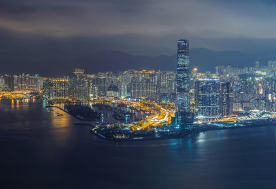 Aerial view of illuminated modern buildings in city by river against cloudy sky