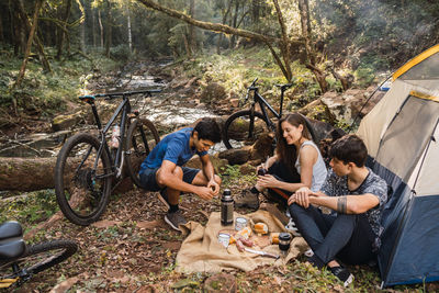 Diverse cyclists sitting on ground near tent and having picnic while hiking together in forest