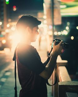 Side view of young man photographing while standing on bridge in city at night