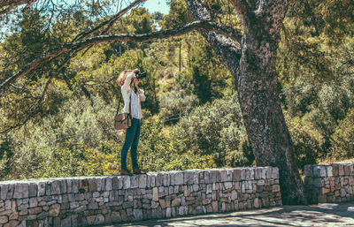 Young woman photographing on stone wall against forest