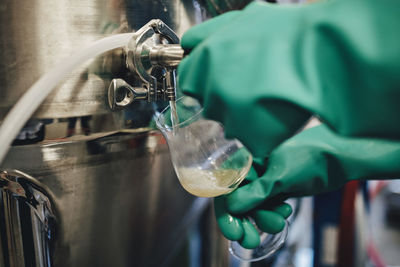 Worker wearing glove pouring beer from container in glass at brewery