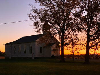 Silhouette tree on field by building against sky at sunset