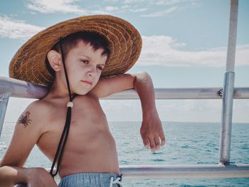 Thoughtful shirtless boy wearing hat while standing by sea against sky