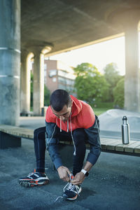 Man tying shoelace while sitting on bench under bridge