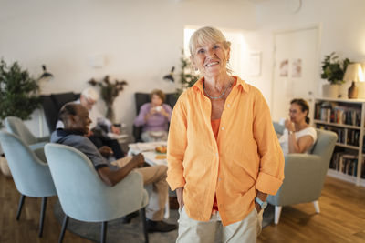Portrait of smiling retired senior woman standing with hands in pockets at nursing home