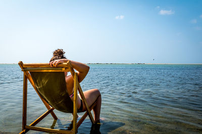 Rear view of young woman sitting on deck chair by sea against sky