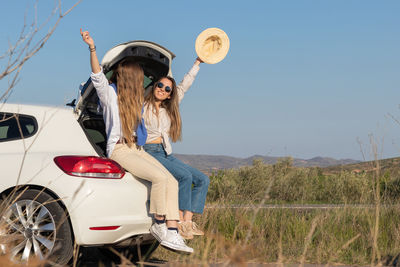 Two friends having good time in spring vacations sitting in back of car while raising arms and hat