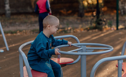 Cute little boy with down syndrome in a funny hat walks in the playground, spinning on a carousel