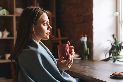 Young woman in cozy grey scarf with mug of tea in hands looks out the window and rests in studio