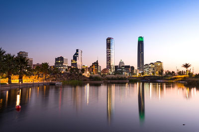 Reflection of illuminated buildings in water against clear blue sky