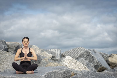 Full length of woman meditating while sitting on rock against cloudy sky