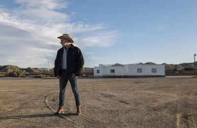 Adult man in cowboy hat standing in front of white building against sky