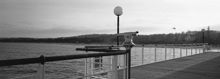 Coin-operated binoculars on railing by lake against sky