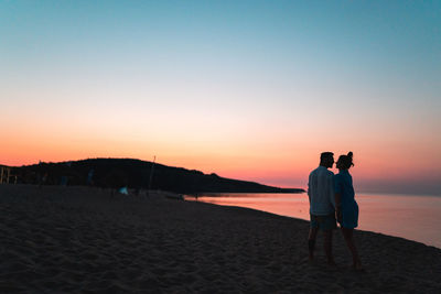 Rear view of men standing on beach against sky during sunset