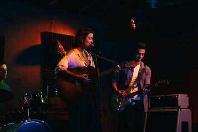 Group of people in casual clothes playing guitars and drums while woman singing and performing song in club with neon lights