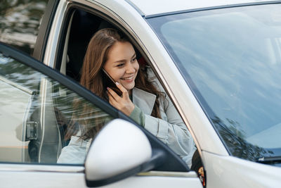 Beautiful girl with long hair in a grey trench coat using smartphone call gets into the car