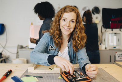 Portrait of smiling redhead female technician sitting at workbench while colleagues working in creative office