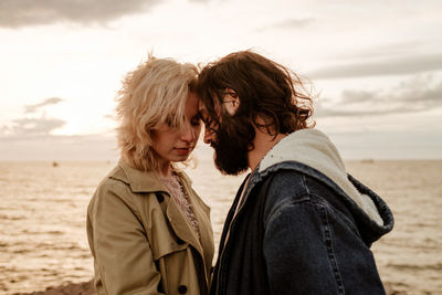 Boyfriend and girlfriend in outerwear touching foreheads against rippling sea and cloudy sky in evening in aviles, spain