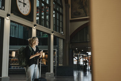 Thoughtful woman with smart phone carrying luggage while standing at station