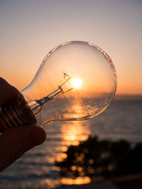 Close-up of hand holding light bulb against sky during sunset